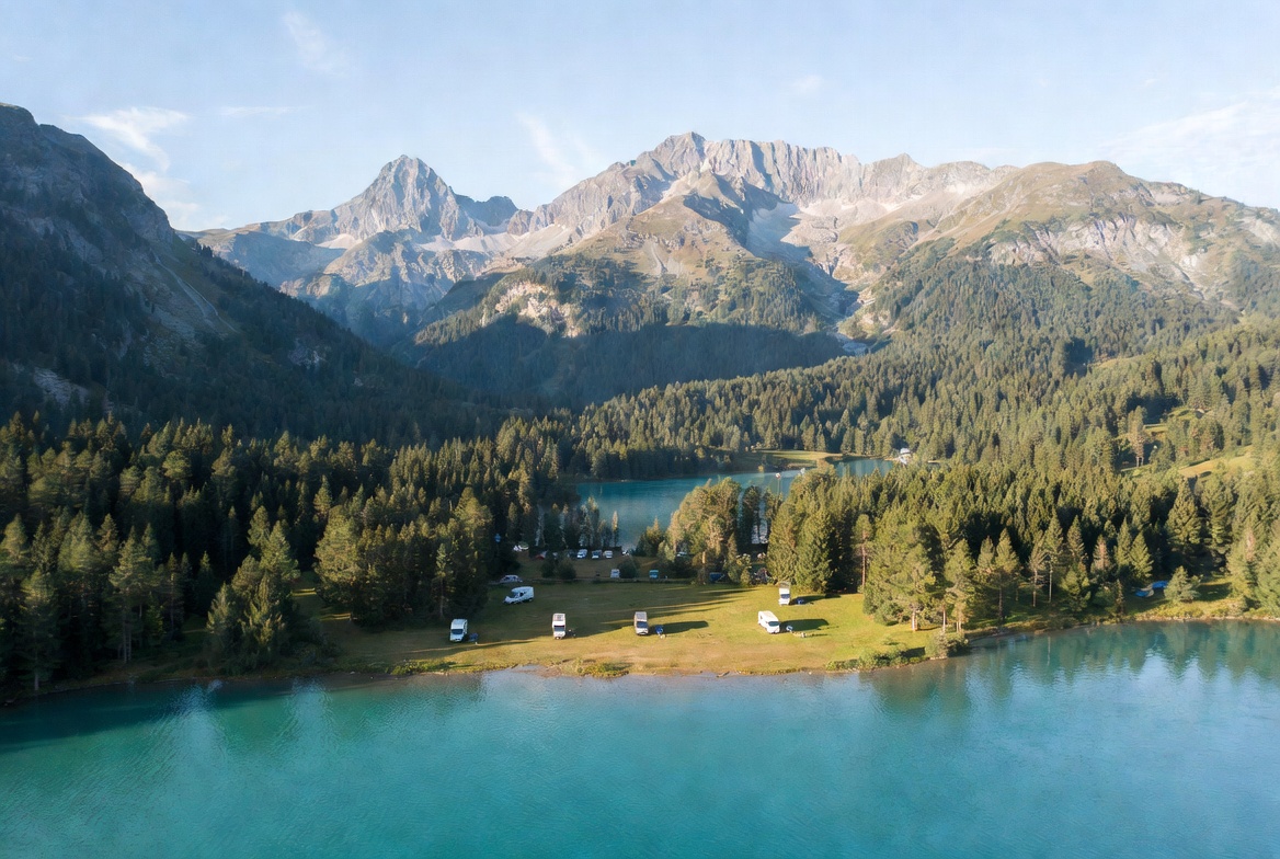Vista aérea de un área de autocaravanas junto a un lago alpino turquesa rodeado de bosque de pinos y montañas nevadas
